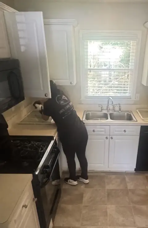 A Santos Cleaning team member in a company shirt wiping out an upper cabinet, with refrigerator drawers washed and drying on the counter